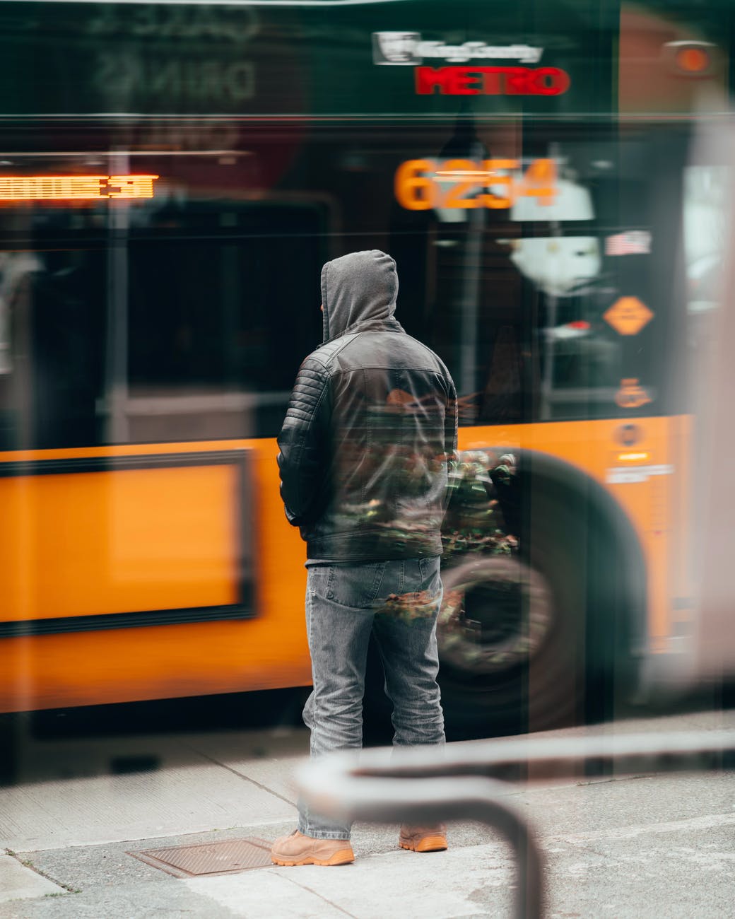 man in black jacket and blue denim jeans standing in front of yellow bus